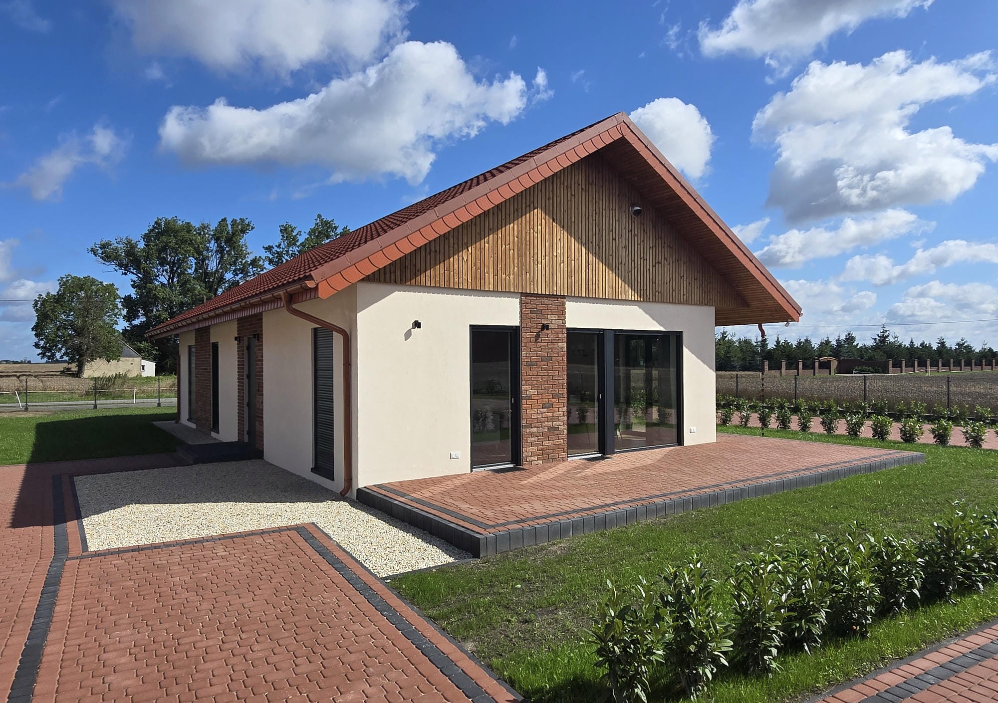 Nowoczesny dom z białymi ścianami, czerwonym dachem i dużymi oknami, otoczony zielonym ogrodem i ścieżkami z cegieł