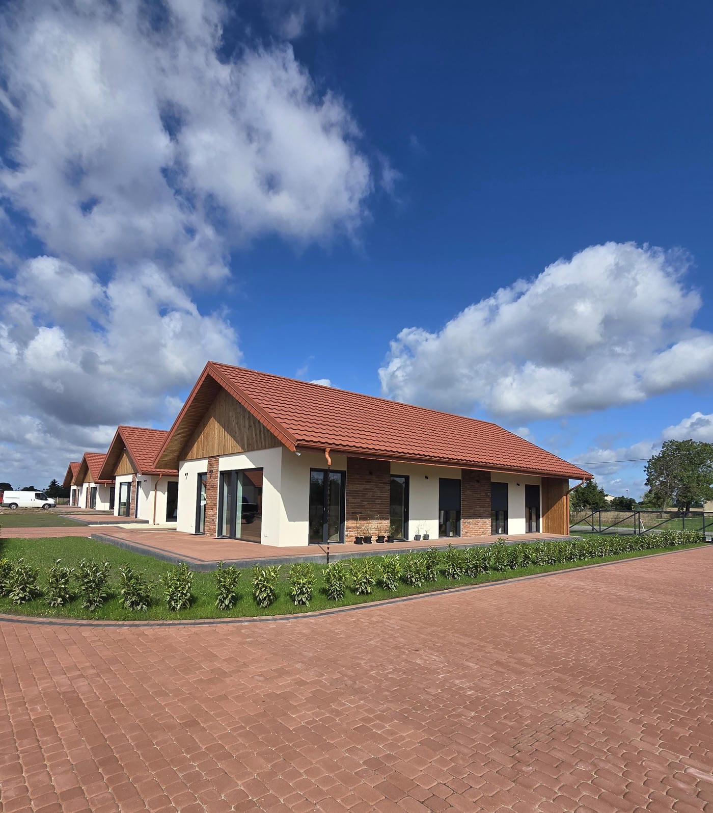 Nowoczesny dom z czerwonym dachem, białymi ścianami i dużymi oknami, otoczony zielonym trawnikiem pod niebieskim niebem z chmurami