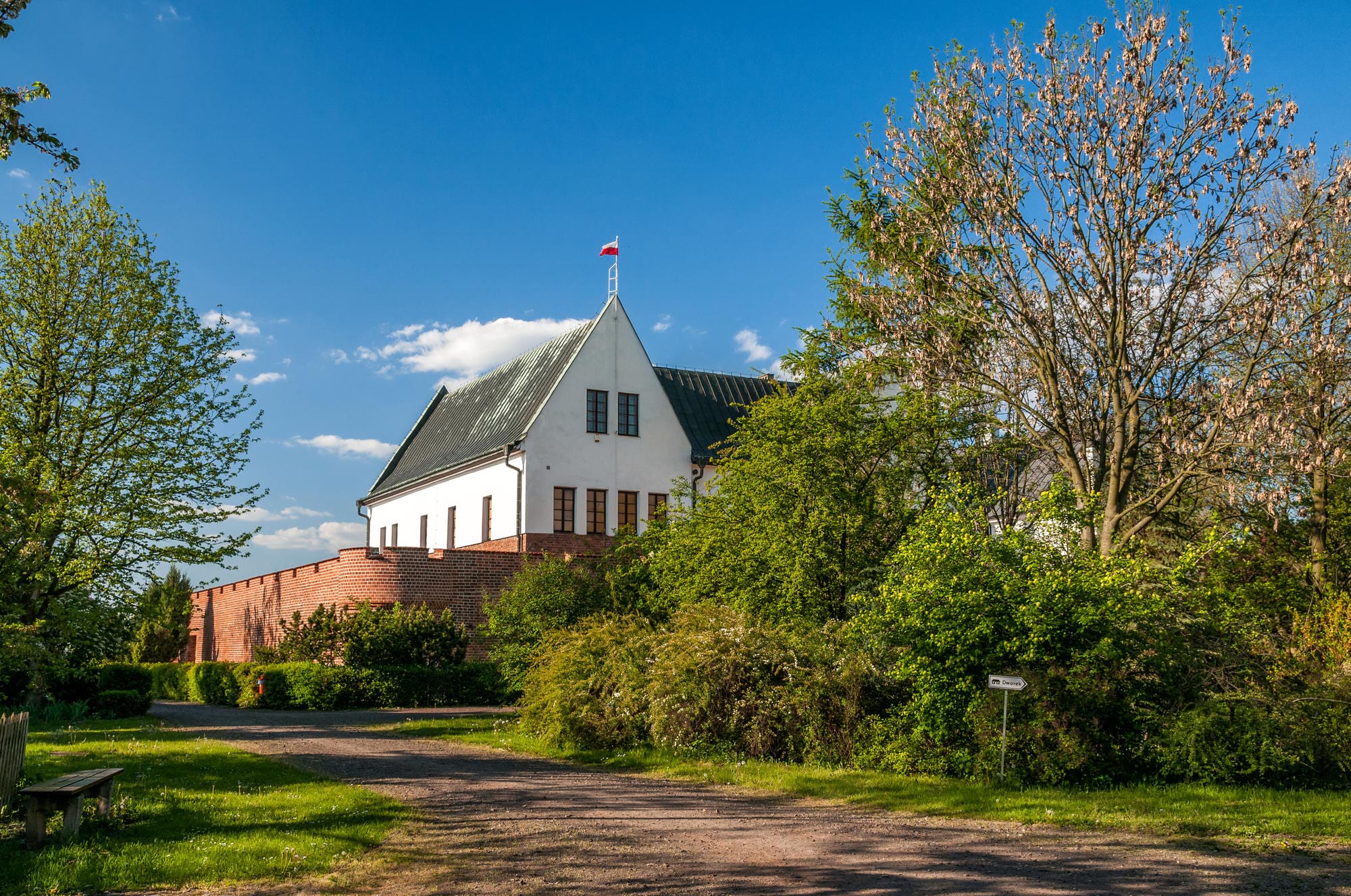 Biały budynek z czarna dachówką na wzniesieniu, otoczony zielonymi drzewami i krzewami, przy ścieżce w słoneczny dzień