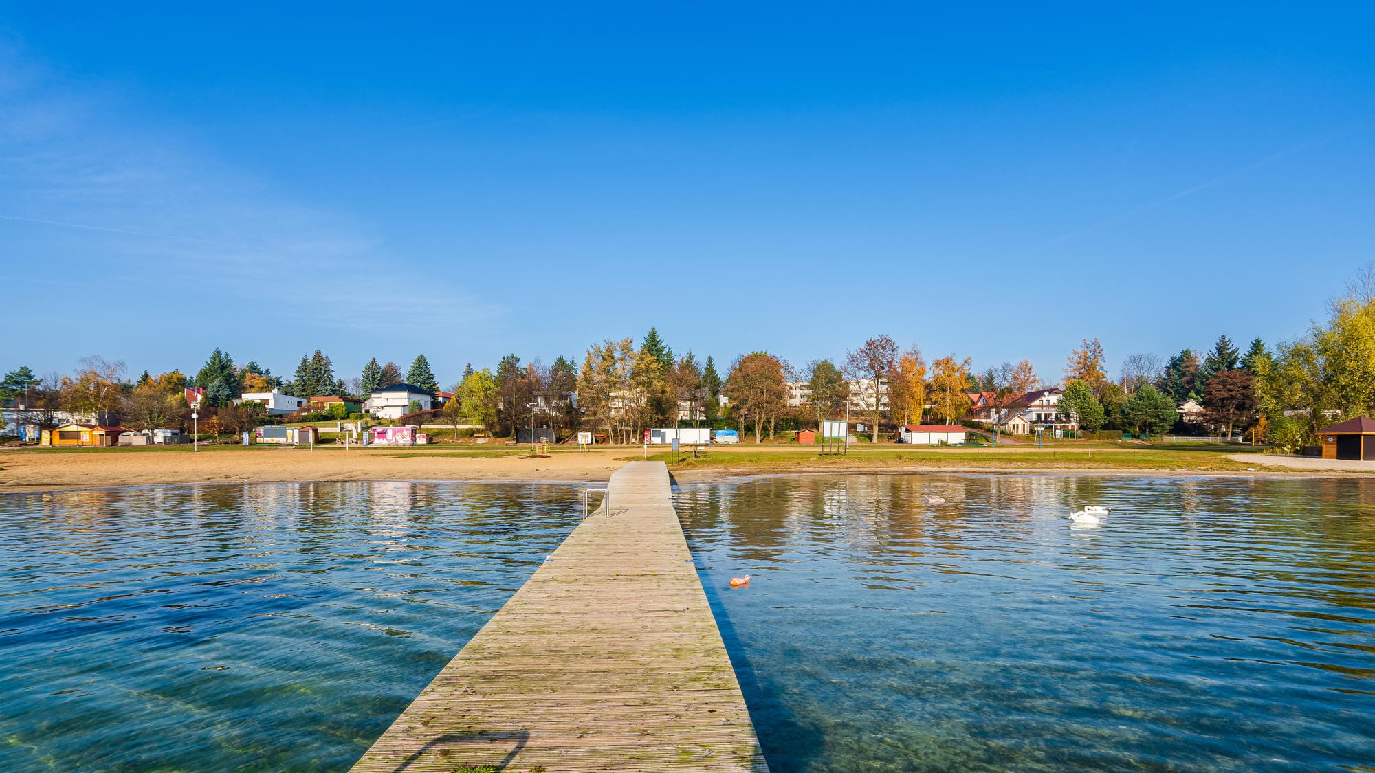 Długa drewniana kładka prowadzi do brzegu jeziora z widokiem na domy i drzewa w jesiennych barwach przy spokojnej wodzie