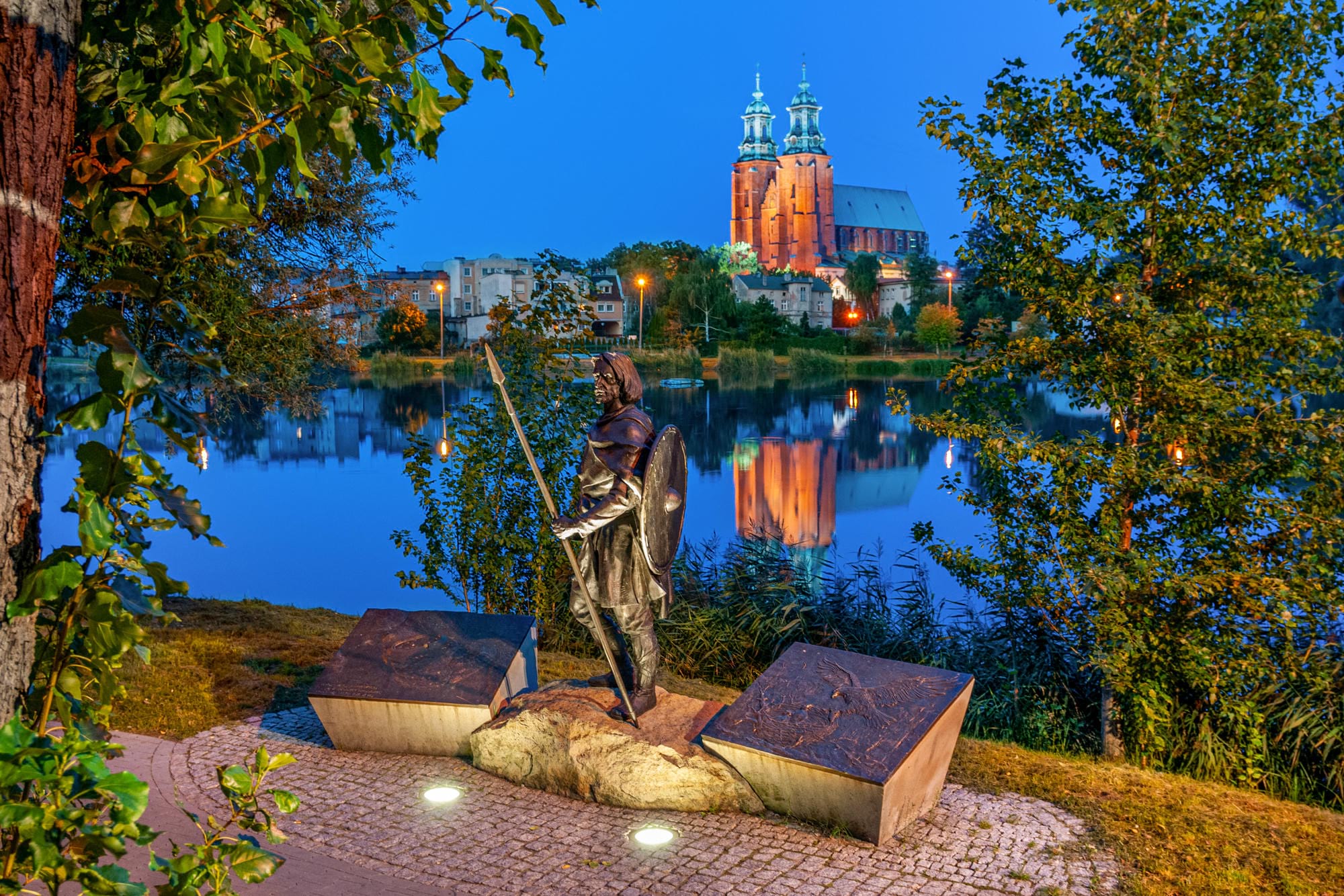 Statua rycerza z tarczą i włócznią na tle jeziora i oświetlonego kościoła w nocy