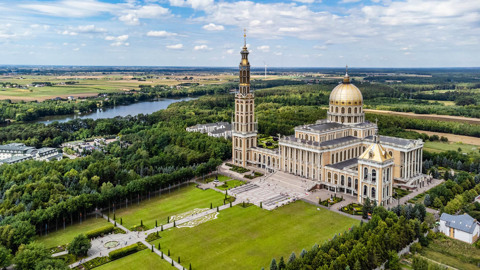 Widok na dużą bazylikę z kopułą i wysoką wieżą, otoczoną zielonymi terenami i rzeką w tle w krajobrazie wiejskim