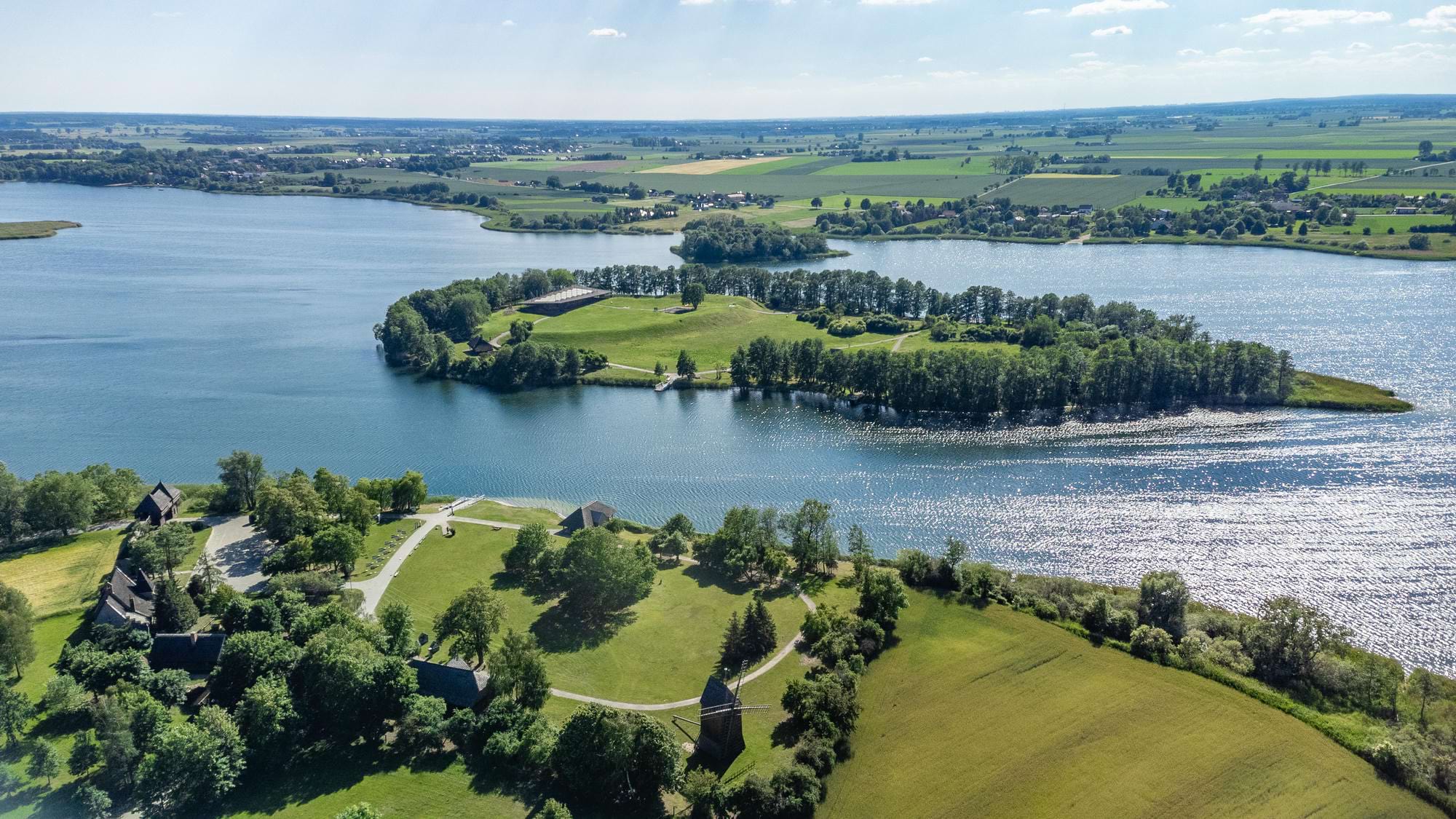 Widok na wyspę z zielonymi terenami, stawem i kilkoma wiatrakami, otoczona wodami i roślinnością, na tle pola i nieba