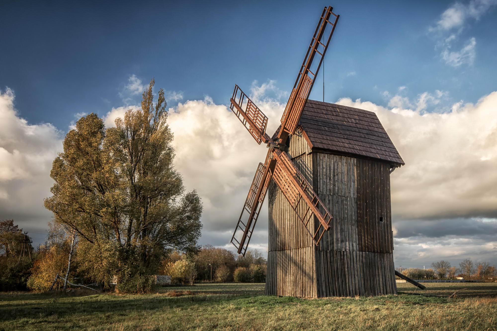 Stary wiatrak z drewnianymi skrzydłami na tle chmurnego nieba i drzew o złotych liściach